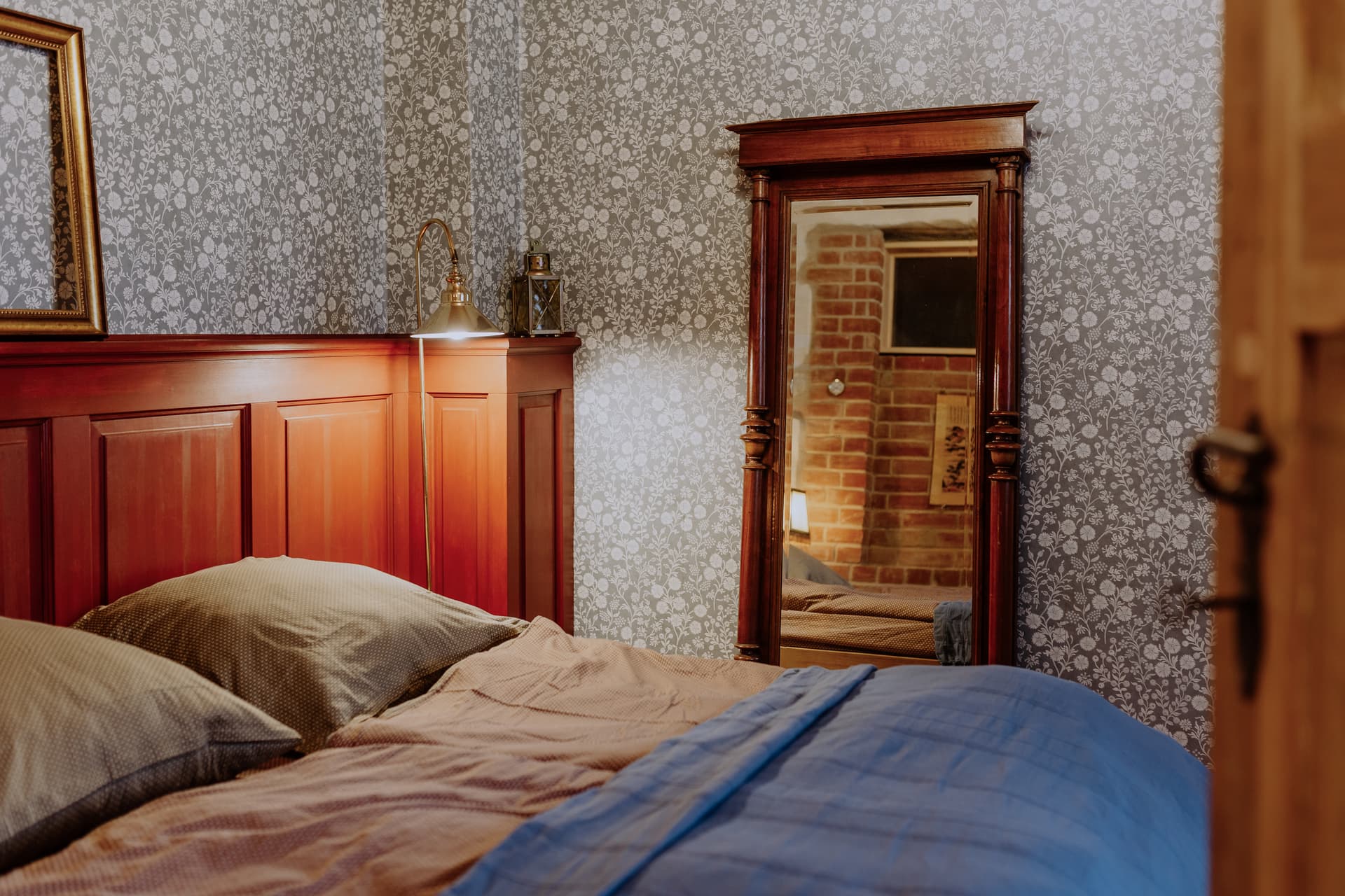 Schlafzimmer 1 mit Doppelbett, rot lackiertem Holzkopfteil, floraler Wandtapete und antikem Standspiegel