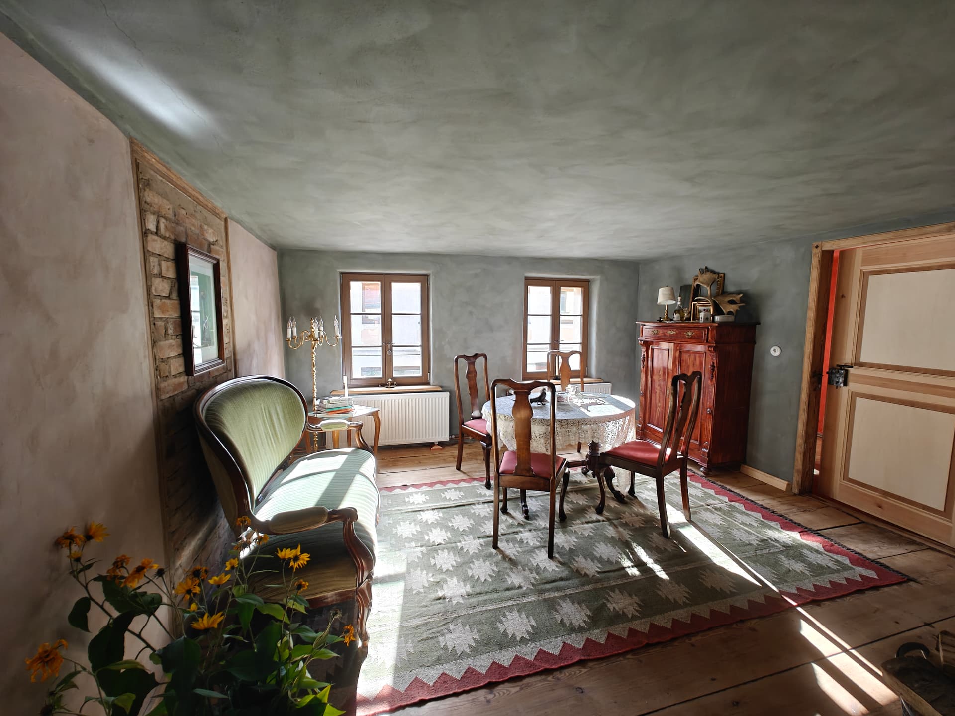Sonniger Wohn- und Speisesalon in Die Stuben mit Biedermeiersofa, runder Tafel mit Spitzendecke, Buffet und Deckenbalken